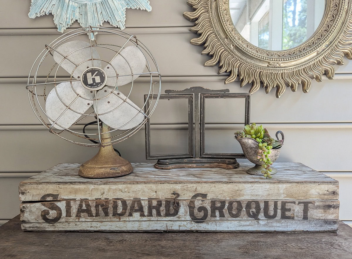 vintage decor on porch including old fan, empty frame, succulents in a gravy boat, and a croquet set box.